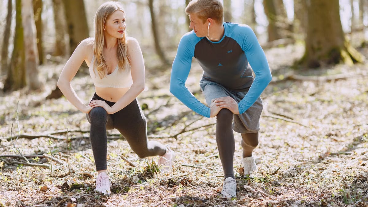 A couple engages in outdoor stretching exercises in a sunlit forest setting, promoting wellness and fitness.